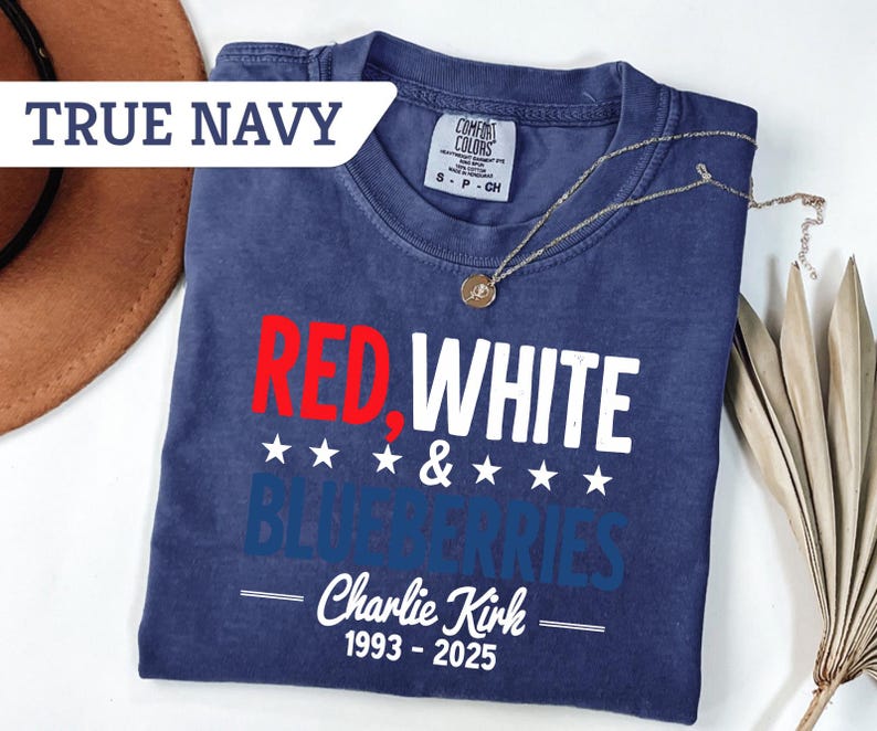 May include: A true navy blue t-shirt with the words "RED, WHITE & BLUEBERRIES" in red, white, and blue. The text "Charlie Kirk 1993-2025" is below the main text. A gold necklace is draped across the shirt.