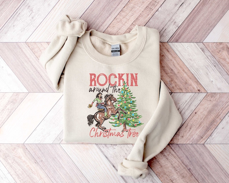 May include: A beige sweatshirt with the text "Rockin' around the Christmas Tree" and a graphic of a person riding a horse in front of a Christmas tree.