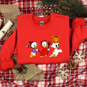 May include: Red sweatshirt featuring a graphic of three cartoon ducks dressed in Halloween costumes. The ducks are wearing a witch hat, devil horns, and a ghost costume with a jack-o-lantern on their head.