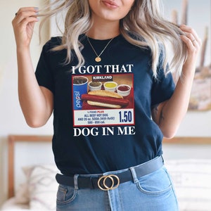 May include: A black t-shirt with a vintage-style advertisement for a Kirkland Signature hot dog meal. The ad features a hot dog, condiments, and a Pepsi can. The text on the shirt reads "I got that DOG in me."