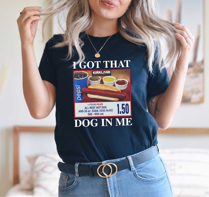 May include: A black t-shirt with a vintage-style advertisement for a Kirkland Signature hot dog meal. The ad features a hot dog, condiments, and a Pepsi can. The text on the shirt reads "I got that DOG in me."