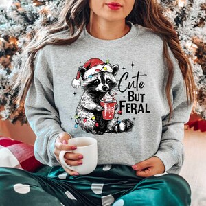 May include: A gray sweatshirt with a cute cartoon raccoon wearing a Santa hat and holding a cup of hot chocolate. The text "Cute But Feral" is printed on the sweatshirt.