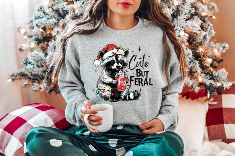 May include: A gray sweatshirt with a cute cartoon raccoon wearing a Santa hat and holding a cup of hot chocolate. The text "Cute But Feral" is printed on the sweatshirt.