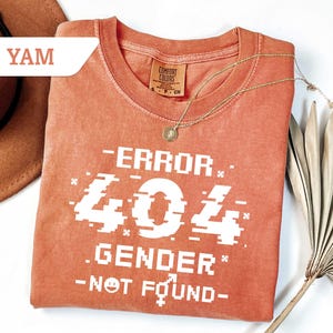May include: A burnt orange t-shirt with a white graphic that says "ERROR 404 GENDER NOT FOUND" with a pixelated font.