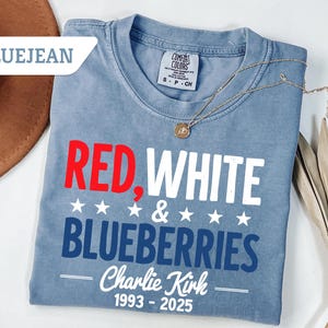 May include: Blue jean colored t-shirt with the words "RED, WHITE & BLUEBERRIES" in red, white, and blue. The shirt also has the text "Charlie Kirk 1993-2025". A gold necklace is draped across the shirt.