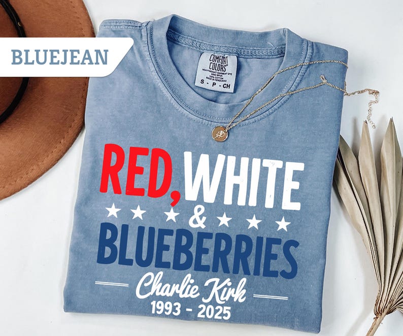 May include: Blue jean colored t-shirt with the words "RED, WHITE & BLUEBERRIES" in red, white, and blue. The shirt also has the text "Charlie Kirk 1993-2025". A gold necklace is draped across the shirt.