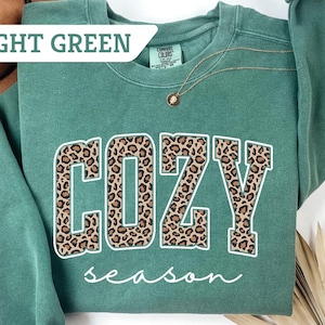 May include: Light green sweatshirt with the words "COZY season" in a white and leopard print design. A gold necklace is visible. The sweatshirt is a Comfort Colors brand.