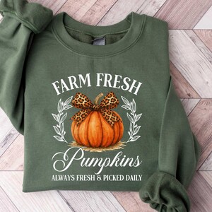 May include: An olive green sweatshirt featuring the text "FARM FRESH Pumpkins" with a pumpkin design. The pumpkin has a leopard print bow and is surrounded by a white wreath. The text "ALWAYS FRESH & PICKED DAILY" is below the word "Pumpkins".