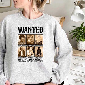 May include: A grey sweatshirt with a graphic print of a wanted poster featuring black and white photographs of notable women. The text on the poster reads "WANTED" and "WELL-BEHAVED WOMEN SELDOM MAKE HISTORY".