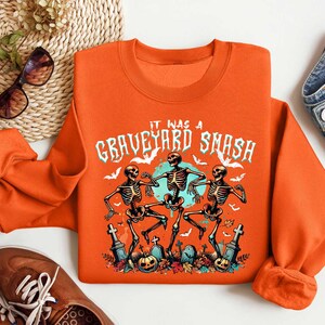 May include: Orange sweatshirt with a graphic of three skeletons dancing and the text "It was a Graveyard Smash".