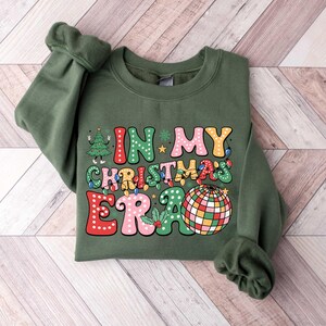 Op de afbeelding: Groen sweatshirt met een feestelijk ontwerp dat "In My Christmas Era" zegt met een discobal en een kerstboom.