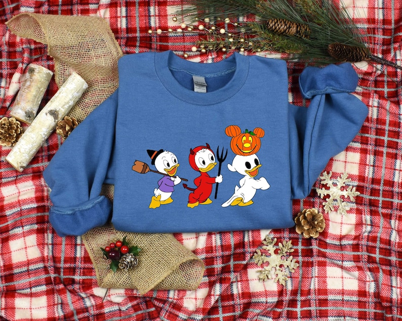 May include: A blue sweatshirt featuring a graphic of three cartoon ducks dressed in Halloween costumes. The ducks are wearing a witch hat, devil horns, and a ghost costume with a pumpkin on its head.
