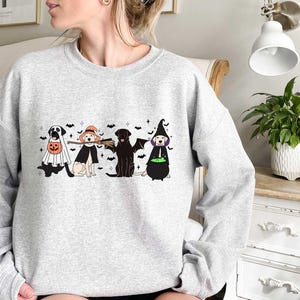 May include: A gray sweatshirt with a Halloween-themed design featuring four dogs dressed in costumes. The dogs are wearing a ghost costume, a witch costume, a broom costume, and a cauldron costume. The design is printed on the front of the sweatshirt.