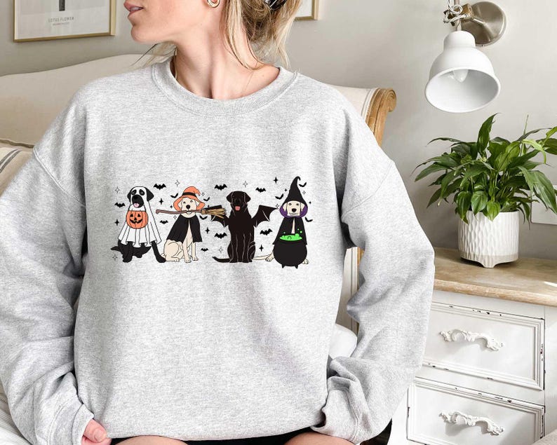 May include: A gray sweatshirt with a Halloween-themed design featuring four dogs dressed in costumes. The dogs are wearing a ghost costume, a witch costume, a broom costume, and a cauldron costume. The design is printed on the front of the sweatshirt.
