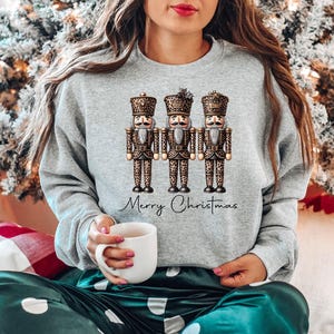 May include: A gray sweatshirt with a graphic of three nutcrackers wearing leopard print outfits. The nutcrackers are holding a banner that says "Merry Christmas".