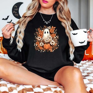 May include: Black sweatshirt with a white ghost in a floral wreath with orange pumpkins. The ghost is surrounded by orange, white, and brown flowers. The sweatshirt has a crew neck and long sleeves.