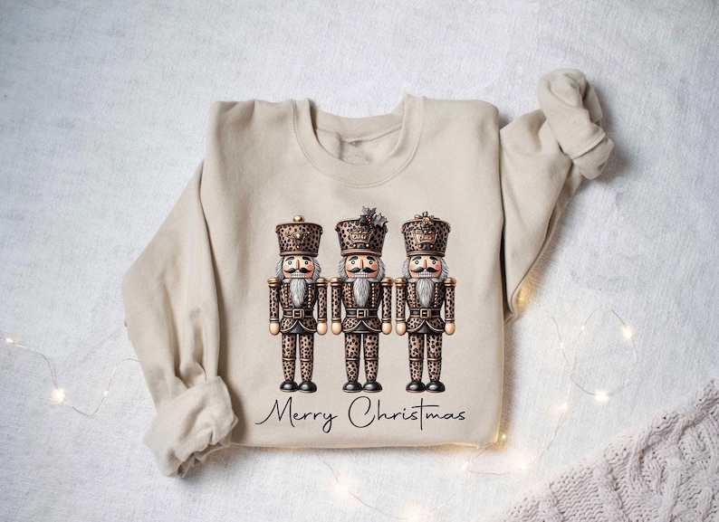 May include: A beige sweatshirt with a graphic of three nutcracker figurines wearing leopard print outfits. The text "Merry Christmas" is printed below the figurines.