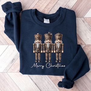 May include: A navy blue sweatshirt with a graphic of three nutcracker figurines wearing gold and brown leopard print outfits. The text "Merry Christmas" is printed below the figurines.