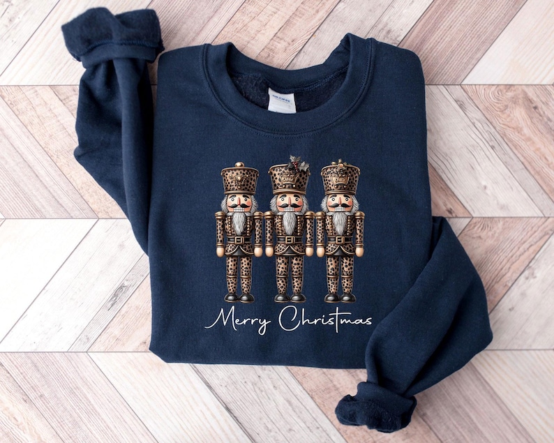 May include: A navy blue sweatshirt with a graphic of three nutcracker figurines wearing gold and brown leopard print outfits. The text "Merry Christmas" is printed below the figurines.