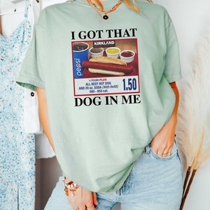May include: A light green t-shirt with the text "I GOT THAT KIRKLAND" and a vintage-style advertisement for a hot dog meal with a Pepsi can, a hot dog, and condiments. The ad text reads "1/4 POUND PLUS ALL BEEF HOT DOG AND 20 oz. SODA (With Refill) 580-850 cal. 1.50 DOG IN ME."