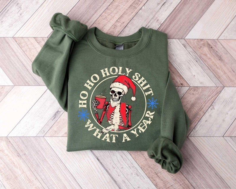 May include: Green sweatshirt with a white circle graphic featuring a skeleton wearing a Santa hat and holding a cup of coffee. The text around the circle reads "Ho Ho Holy Shit What A Year".
