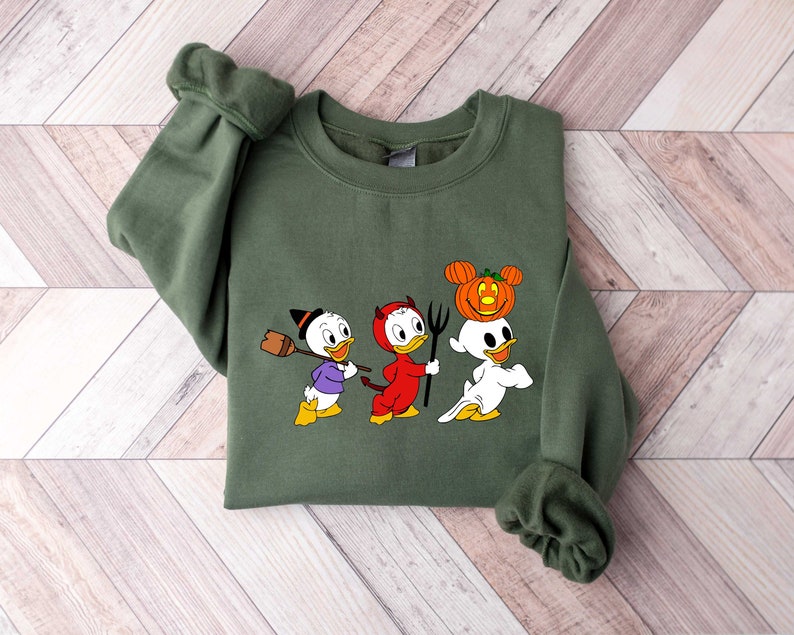 May include: A green sweatshirt with a Halloween-themed design featuring three cartoon ducks dressed as a witch, devil, and ghost. The ghost duck is wearing a pumpkin on its head.