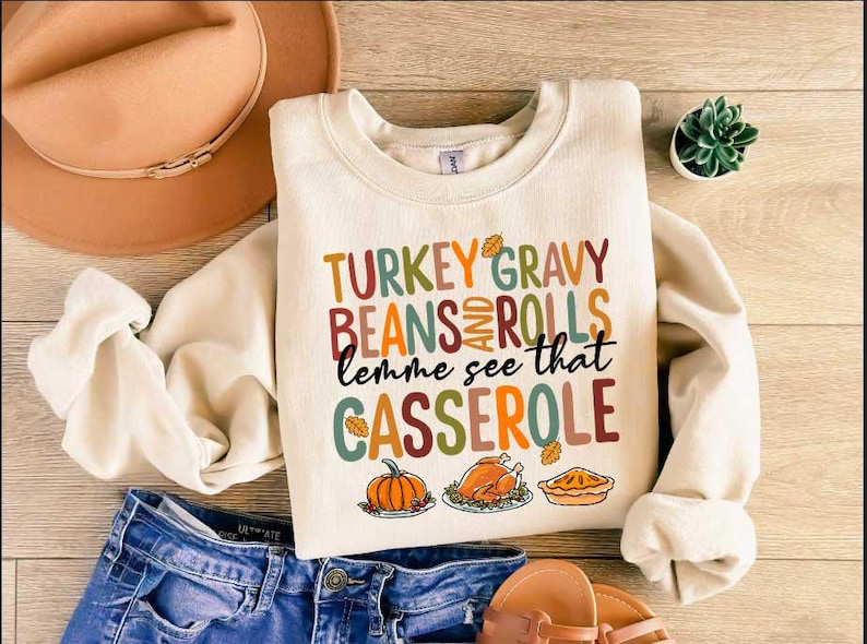 May include: A cream-colored sweatshirt with the text "TURKEY GRAVY BEANS AND ROLLS lemme see that CASSEROLE" in colorful letters. The sweatshirt has a fall-themed design with images of a pumpkin, a turkey, and a pie.