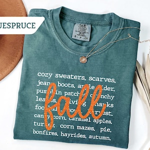 May include: A teal Comfort Colors t-shirt with the word "fall" in orange glitter script. The shirt lists fall-themed words like "sweaters," "boots," and "hayrides." A gold necklace and a brown hat are also visible.