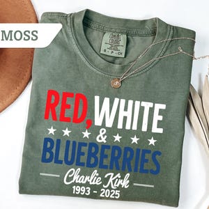 May include: A moss green t-shirt with the words "RED, WHITE & BLUEBERRIES" in red, white, and blue. The text "Charlie Kirk 1993-2025" is printed below. A gold necklace and a brown hat are also visible.