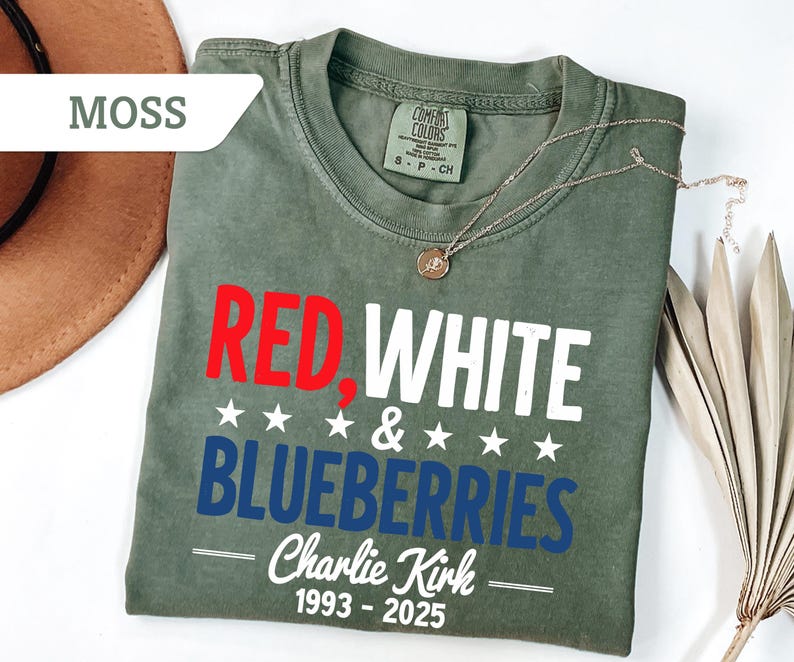 May include: A moss green t-shirt with the words "RED, WHITE & BLUEBERRIES" in red, white, and blue. The text "Charlie Kirk 1993-2025" is printed below. A gold necklace and a brown hat are also visible.