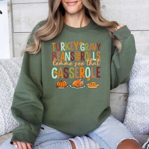 May include: Green sweatshirt with the text "TURKEY GRAVY BEANS AND ROLLS lemme see that CASSEROLE" in a festive font. The text is surrounded by images of a pumpkin, a turkey, and a casserole dish.