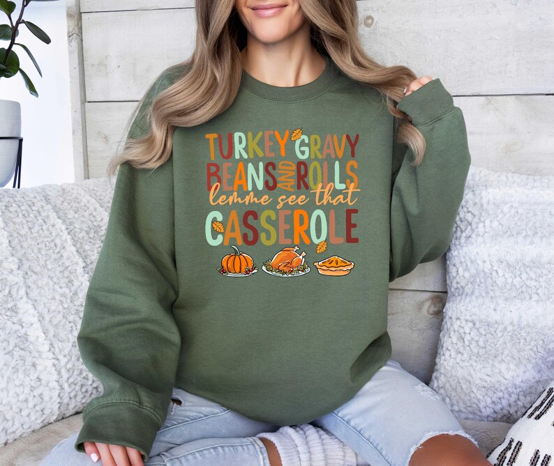 May include: Green sweatshirt with the text "TURKEY GRAVY BEANS AND ROLLS lemme see that CASSEROLE" in a festive font. The text is surrounded by images of a pumpkin, a turkey, and a casserole dish.