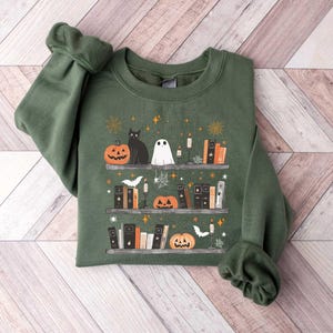 May include: A green sweatshirt with a Halloween-themed design featuring a black cat, a ghost, pumpkins, and books on a shelf. The design is surrounded by stars and other festive elements.