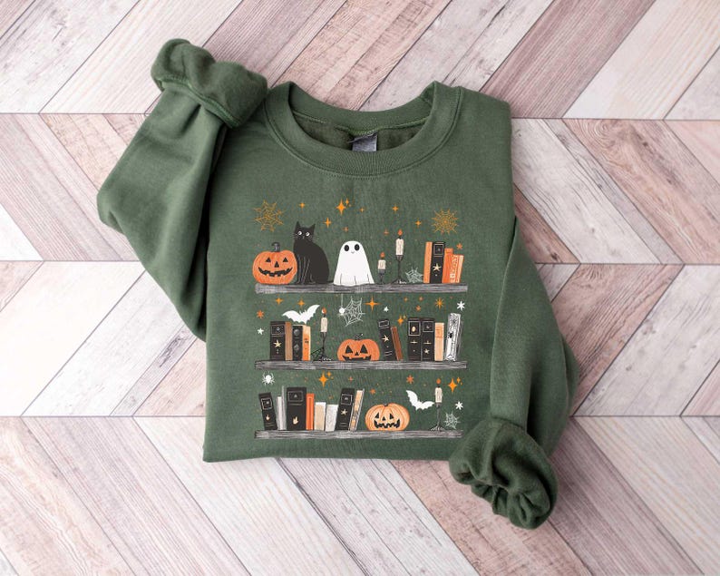 May include: A green sweatshirt with a Halloween-themed design featuring a black cat, a ghost, pumpkins, and books on a shelf. The design is surrounded by stars and other festive elements.