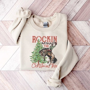 May include: A beige sweatshirt with a vintage-style graphic of a cowboy riding a horse around a Christmas tree. The text reads "Rockin' around the Christmas Tree".