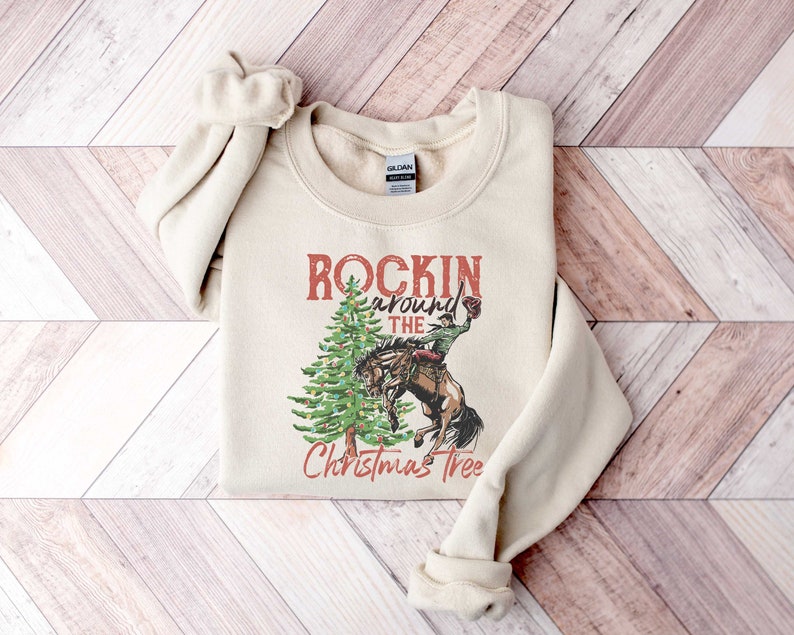 May include: A beige sweatshirt with a vintage-style graphic of a cowboy riding a horse around a Christmas tree. The text reads "Rockin' around the Christmas Tree".