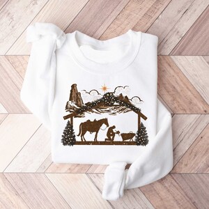 Puede incluir: Sudadera blanca con una silueta marr&oacute;n de un pesebre, que incluye un caballo, figuras y una estrella. El dise&ntilde;o presenta un establo, monta&ntilde;as y &aacute;rboles de Navidad, creando un tema navide&ntilde;o r&uacute;stico. La sudadera est&aacute; hecha de un material suave.