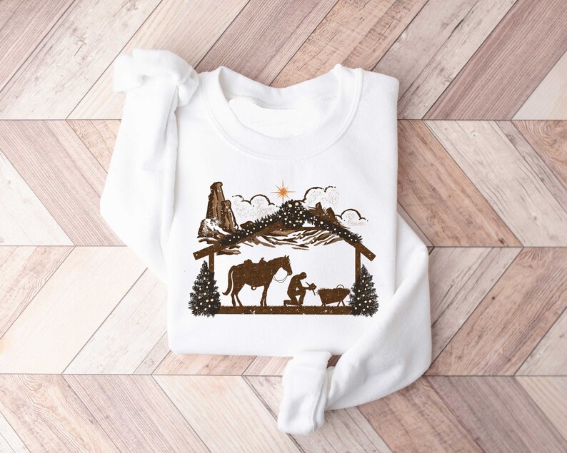 Puede incluir: Sudadera blanca con una silueta marr&oacute;n de un pesebre, que incluye un caballo, figuras y una estrella. El dise&ntilde;o presenta un establo, monta&ntilde;as y &aacute;rboles de Navidad, creando un tema navide&ntilde;o r&uacute;stico. La sudadera est&aacute; hecha de un material suave.