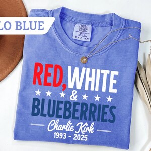 May include: A blue t-shirt with the words "RED, WHITE & BLUEBERRIES" in red and white text. The shirt also has the text "Charlie Kirk 1993-2025". A gold necklace and a brown hat are also in the image.