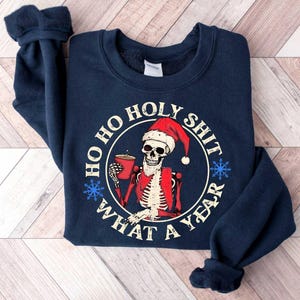 May include: Navy blue sweatshirt with a white skull wearing a Santa hat holding a cup of coffee. The text on the sweatshirt reads "Ho Ho Holy Shit What A Year".