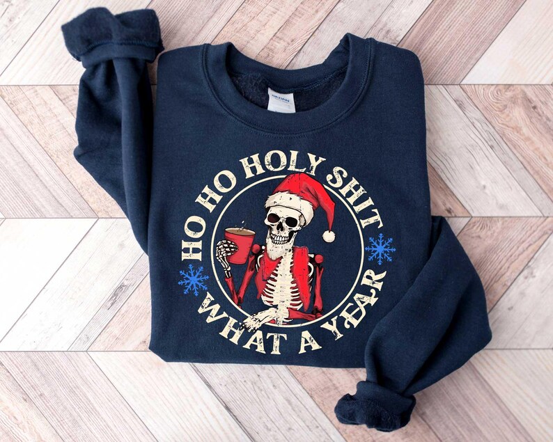 May include: Navy blue sweatshirt with a white skull wearing a Santa hat holding a cup of coffee. The text on the sweatshirt reads "Ho Ho Holy Shit What A Year".