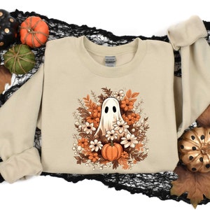 May include: A beige sweatshirt with a white ghost surrounded by orange and white flowers. The ghost has a single eye and a small mouth. The flowers are arranged in a circle around the ghost.