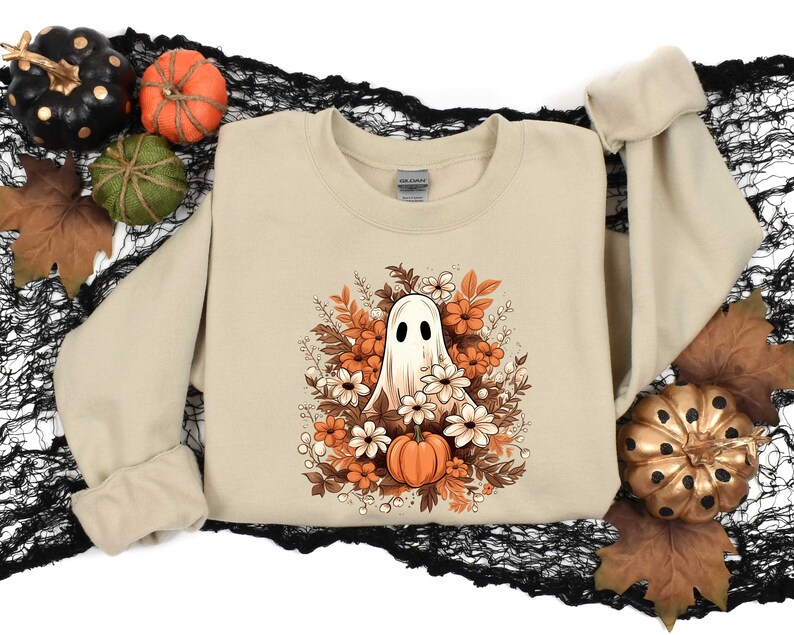 May include: A beige sweatshirt with a white ghost surrounded by orange and white flowers. The ghost has a single eye and a small mouth. The flowers are arranged in a circle around the ghost.