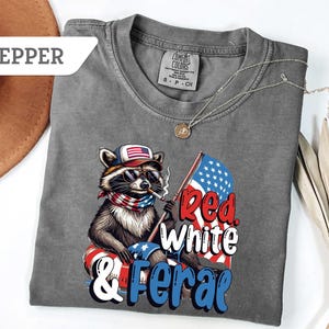 May include: A gray Comfort Colors t-shirt featuring a raccoon wearing sunglasses, a patriotic hat, and a bandana. The raccoon is smoking a cigarette and holding an American flag. The text "Red, White & Feral" is printed on the shirt.