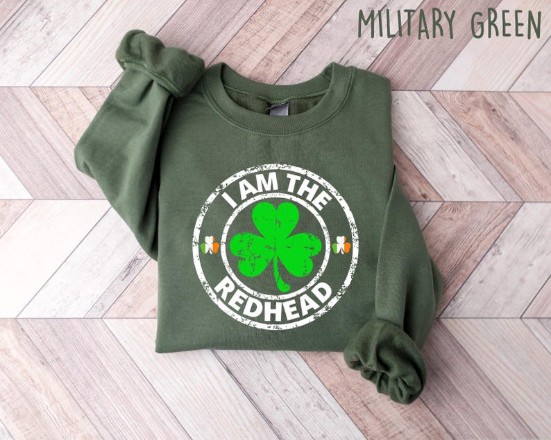 Puede incluir: Una sudadera verde con el texto "I am the redhead" en un c&iacute;rculo blanco con un tr&eacute;bol verde en su interior. El tr&eacute;bol tiene un contorno blanco y un centro verde.