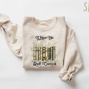 K&ouml;nnte beinhalten: Ein beiger Pullover mit dem Aufdruck "I Have No Shelf Control" auf der Vorderseite. Der Text ist von einer Grafik mit einem Stapel B&uuml;cher mit floralen Akzenten umgeben.