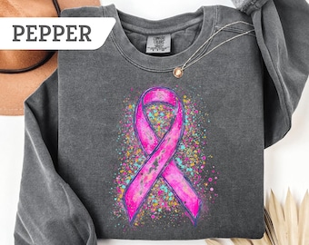 May include: A dark gray sweatshirt with a pink ribbon design and colorful speckles. The word "PEPPER" is in a white box in the upper left corner. A silver necklace with a pendant is visible.