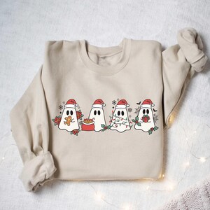 Op de afbeelding: Een beige sweatshirt met een kerstthema met vier geesten die een kerstmanmuts dragen. De geesten houden verschillende kerstspullen vast, waaronder een peperkoek, een cadeau, een lichtjesketting en een presentje.