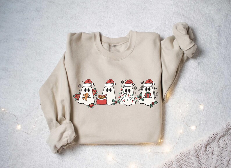 Op de afbeelding: Een beige sweatshirt met een kerstthema met vier geesten die een kerstmanmuts dragen. De geesten houden verschillende kerstspullen vast, waaronder een peperkoek, een cadeau, een lichtjesketting en een presentje.