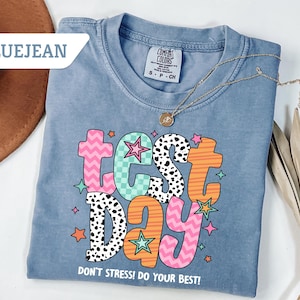 May include: Bluejean Comfort Colors t-shirt with the words "TEST DAY" in colorful, playful lettering. The shirt also has the text "DON'T STRESS! DO YOUR BEST!" and is adorned with star graphics. A gold necklace is draped across the shirt.